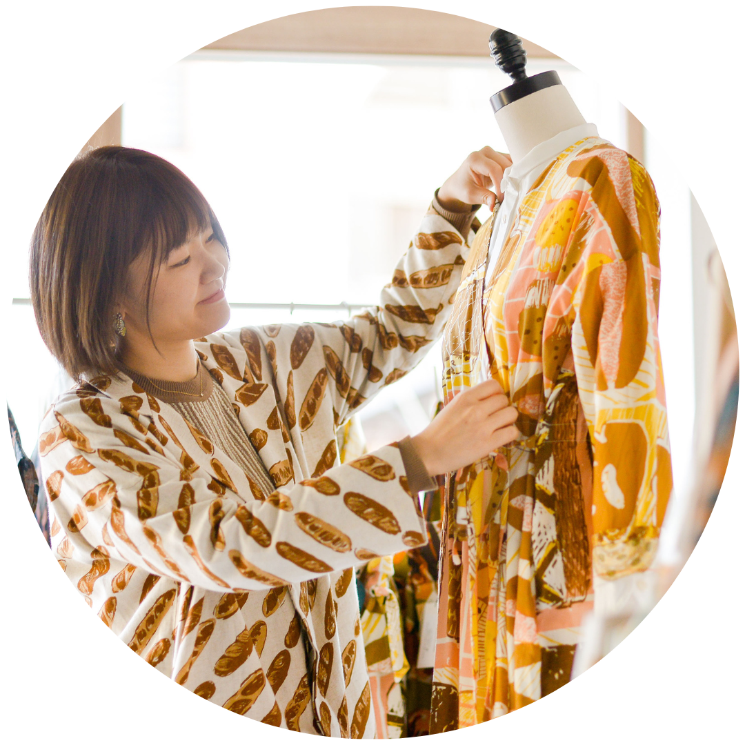 Ako adjusting a colorful dress on a mannequin, surrounded by her fashion designs for Gochisou.